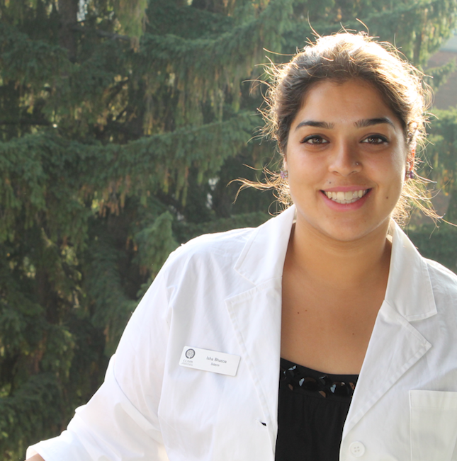 Dr. Bhatoa smiling in a white lab coat in front of evergreen trees